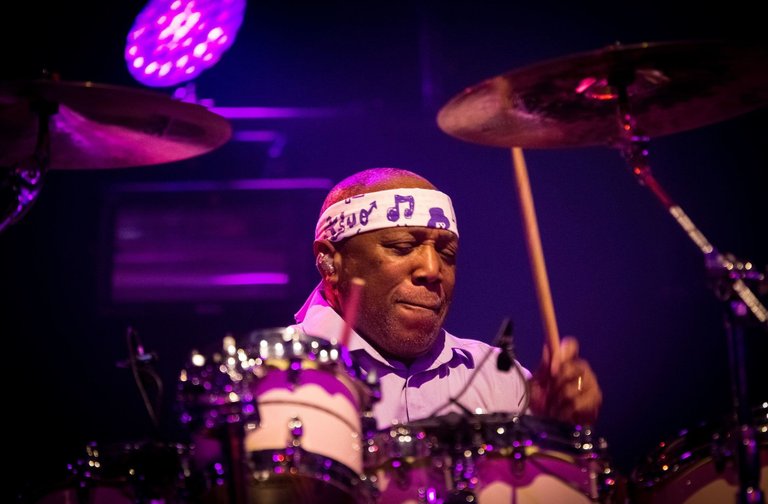 Drummer Billy Cobham plays intensely behind a drum kit on a stage lit with purple and pink lights. He wears a white headband with musical notes and holds drumsticks in motion. The image conveys energy, rhythm, and a live performance atmosphere.