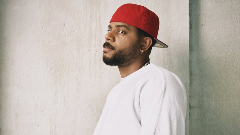 A person with a beard and a red cap stands thoughtfully against a grey wall wearing a white garment.