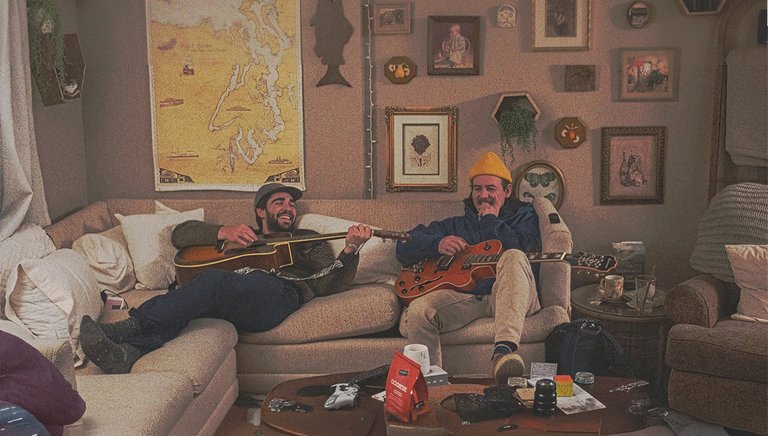 Two men are sitting comfortably on a couch playing acoustic and electric guitars in a room decorated with various ornaments and maps on the walls.