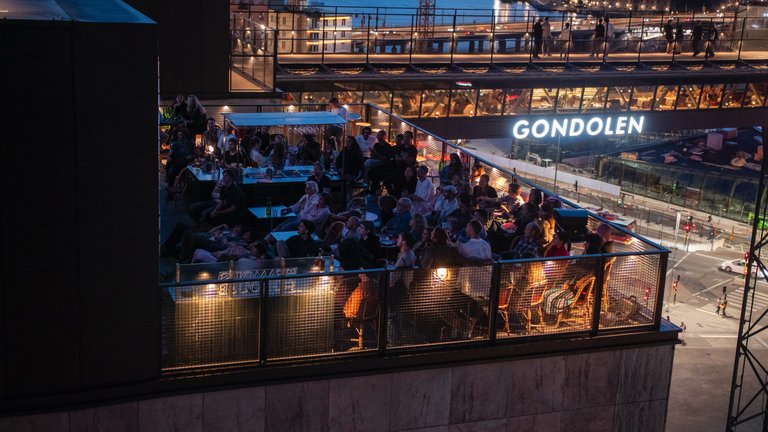 Evening scene on a rooftop terrace at Slussen in Stockholm, where guests gather with views of city lights and the Gondolen building in the background.
