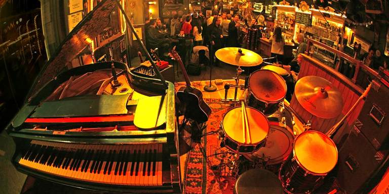The stage at Stampen, a classic jazz and blues bar in Old Town, in Stockholm. The picture is taken from the stage. There’s a drum set, guitar and piano on stage, facing the crowd hanging around the cozy bar.