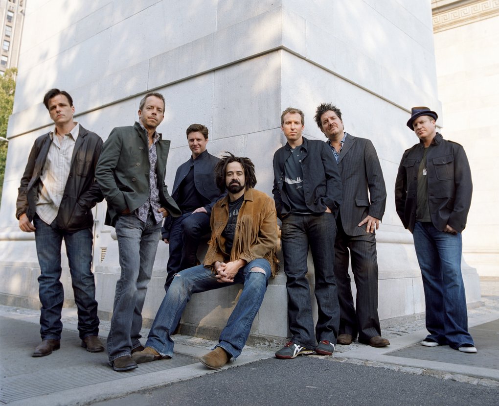 Counting Crows stand and sit leaning against a large white stone building outdoors. They wear a mix of casual and semi-formal clothing. The group poses in a relaxed line.