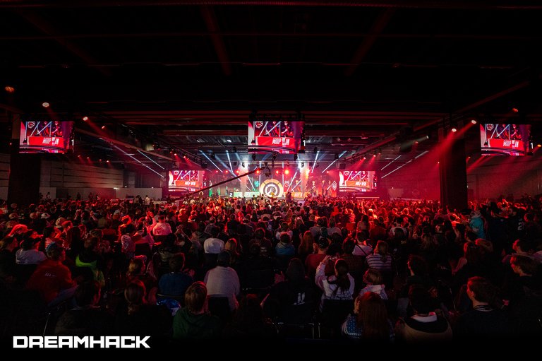 A sea of people in front of a brightly lit stage in a huge, dimly lit hall.