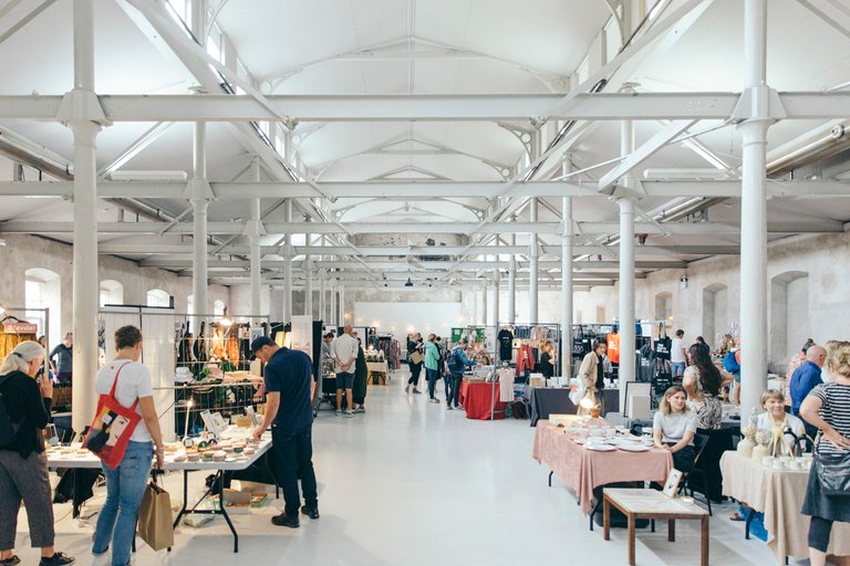 People browsing and selling handmade crafts, clothes, and design items inside a bright industrial hall with high ceilings and metal beams.