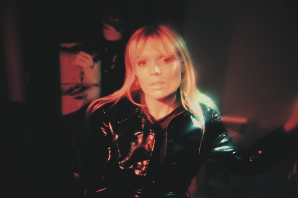 A blonde woman wearing a black leather jacket in a dimly lit room.