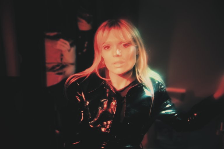 A blonde woman wearing a black leather jacket in a dimly lit room.