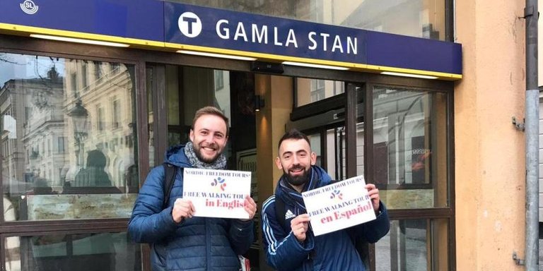 Two guides standing at the entrance of a subway station and holding a sign with the information about their tours.