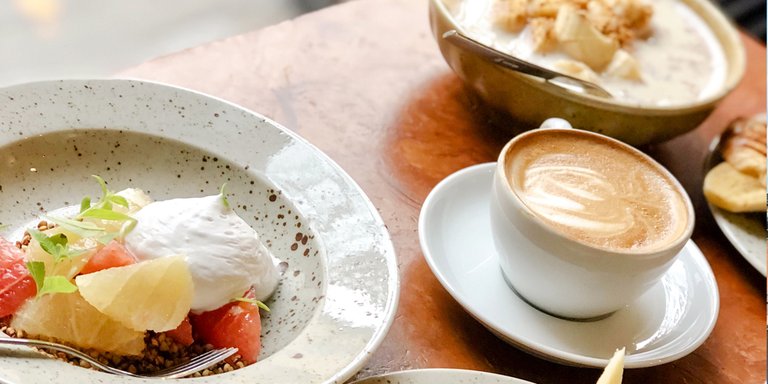 Breakfast at Pom & Flora in Stockholm featuring a bowl of yogurt topped with citrus and fruit and a cup of cappuccino on a wooden table.