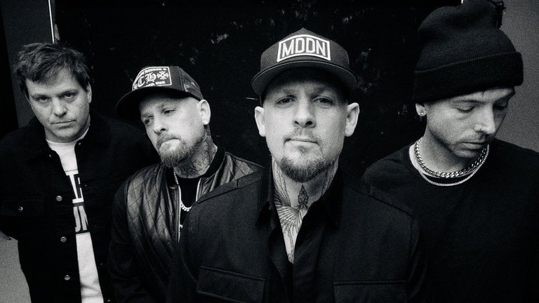 Four men pose in a black-and-white photo, with two wearing caps and all dressed in dark-toned jackets with varied styles.