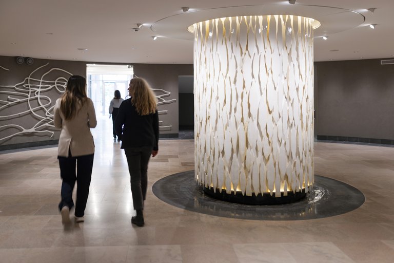 Two people are walking through a modern indoor space with a large illuminated sculpture in the center made of vertical, translucent elements.