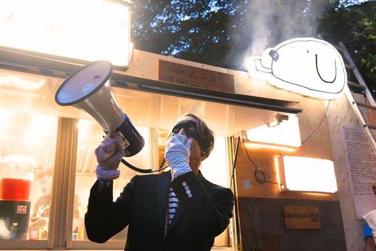 Thomas Öberg, wearing a black jacket, striped shirt, and eye mask, holds a megaphone to his face. In the background, there is a lit-up food truck with Bob Hunds logo on.