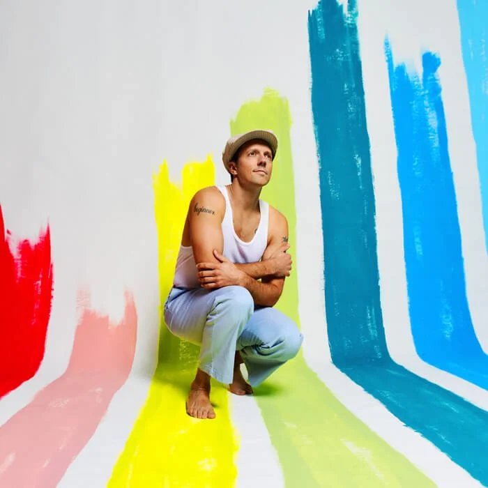 Jason Mraz, in a white tank top and light blue pants crouches barefoot, surrounded by wide paint streaks in red, pink, yellow, green, turquoise, and blue flowing from wall to floor. He wears a cap and looks thoughtfully upward. The background is white with bold color contrasts.