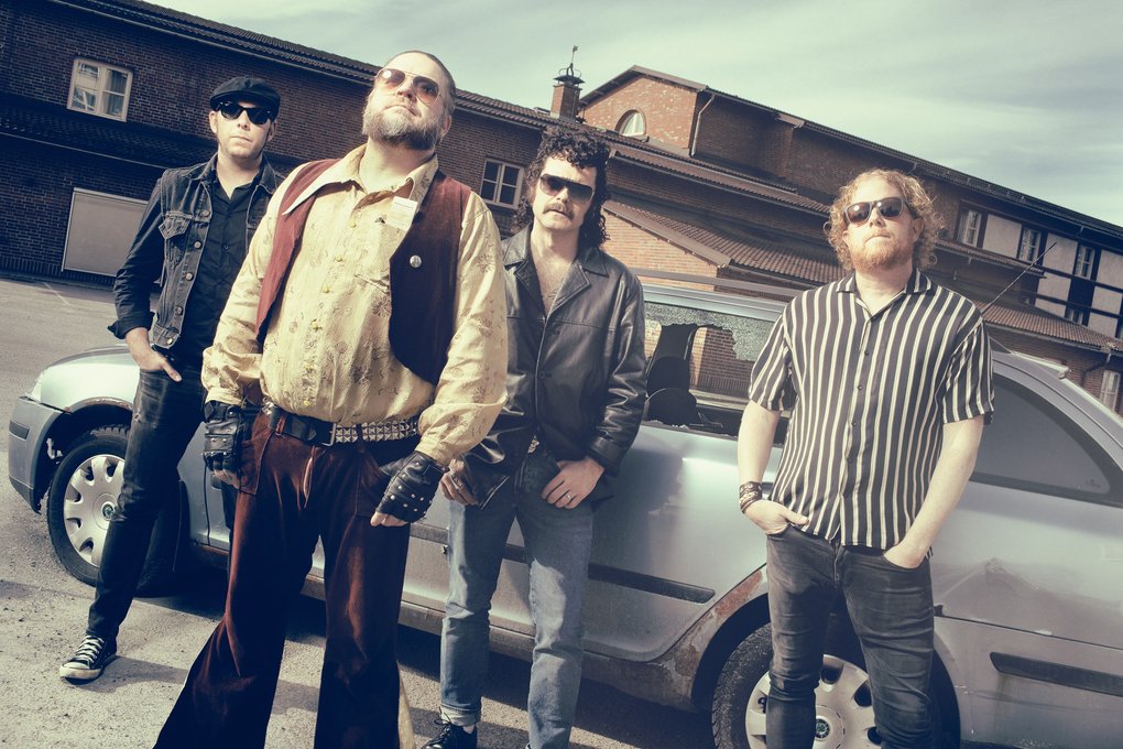 The four members of Korslagda wearing sunglasses and posing next to a rusty, silver colored station wagon.