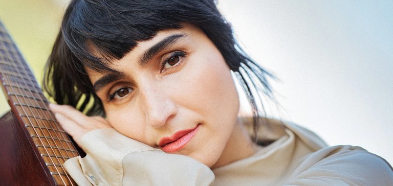 A person with short hair rests their hand on an acoustic guitar and poses with a calm expression.