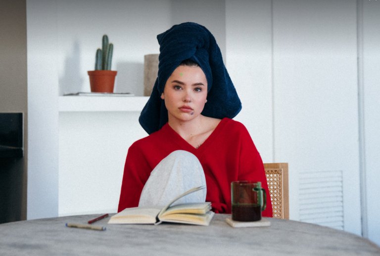 Lauren Spencer Smith sits at a table with an open book and a cup of tea in front of her. She is wearing a red sweater, has a dark blue towel wrapped around her hair, and a small green sticker on her forehead. In the background, a small potted cactus is visible on a shelf.