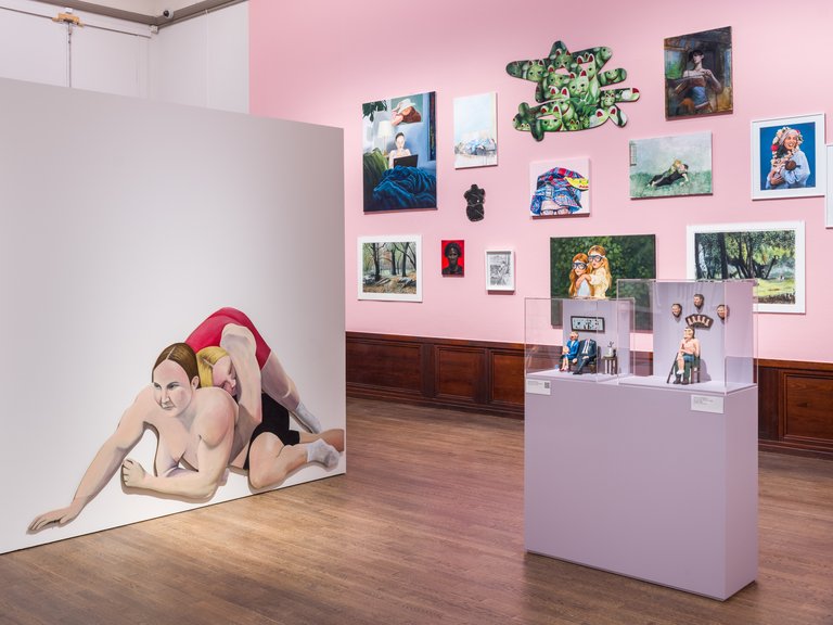 A gallery with pink walls displays a collection of paintings in various sizes along with two sculptures in glass cases on pedestals. On a freestanding wall, a large painting depicts two intertwined bodies in an intimate wrestling pose. The image conveys contemporary art, varied expressions, and a curated exhibition setting.