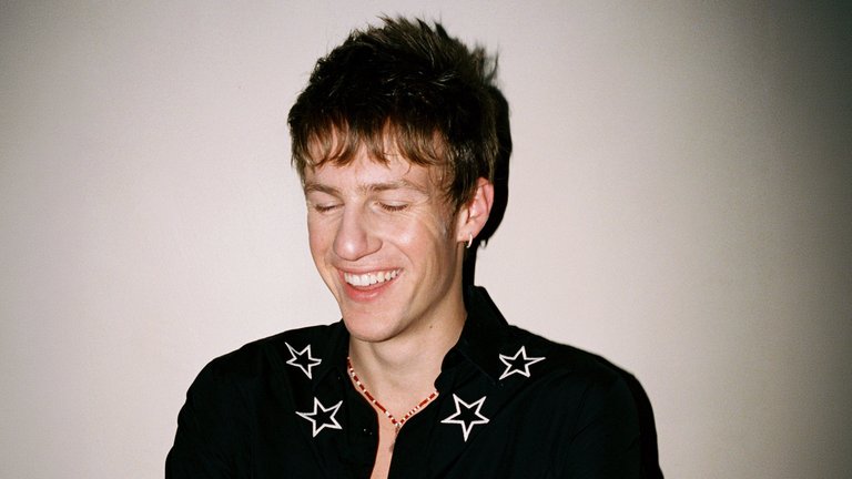 A young man with short, spiky hair is smiling slightly to the side, wearing a black shirt with white star symbols on the shoulders.
