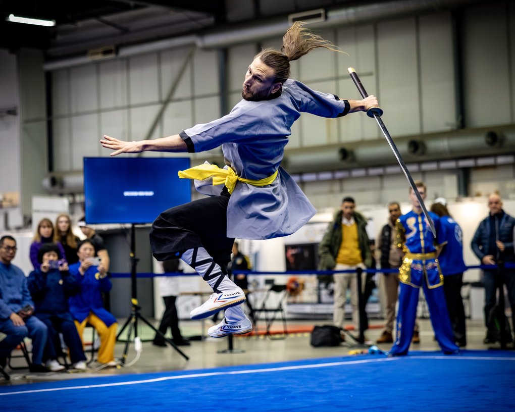 A martial artist in a gray outfit leaps midair with a sword over a blue mat as spectators watch in an indoor arena.