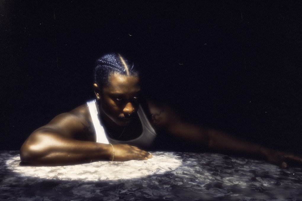 A man in a white undershirt and tattoos on his shoulders leaning on a marble surface under a spotlight.
