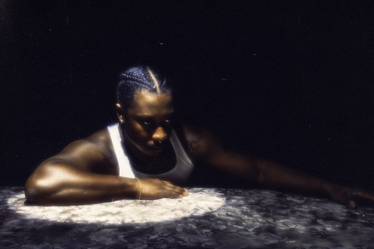 A man in a white undershirt and tattoos on his shoulders leaning on a marble surface under a spotlight.