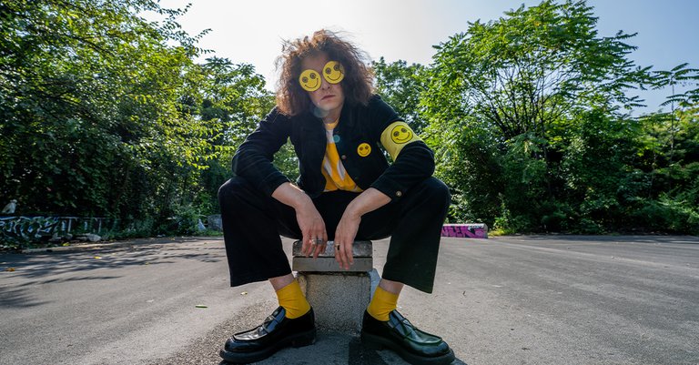 Musician Moon Walker (Harry Springer) sitting on a concrete block outdoors, wearing yellow smiley face glasses and a black jacket