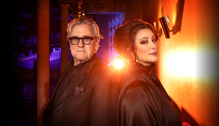 Tommy Körberg and Lisa Nilsson stand back-to-back in dramatic lighting. Körberg wears black clothing and glasses, while Nilsson has styled hair and a black outfit. A strong backlight creates a theatrical and atmospheric effect.
