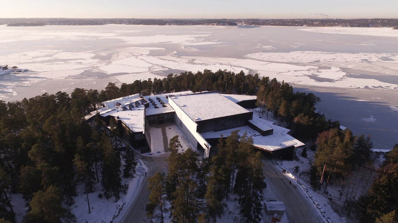 Aerial view of a large building surrounded by trees and water with snow on the ground.