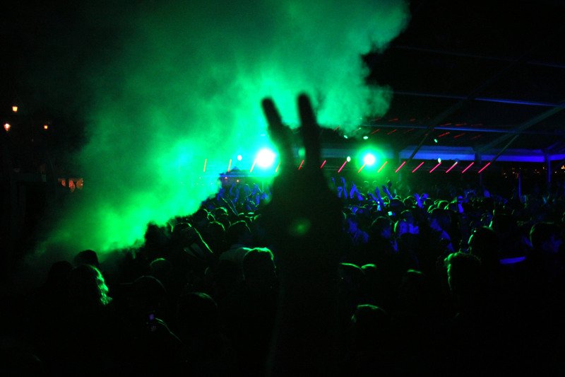 A dense concert crowd with raised hands in the dark, lit by bright green light and smoke. The atmosphere feels intense and energetic, conveying a sense of euphoria and shared excitement among the audience.