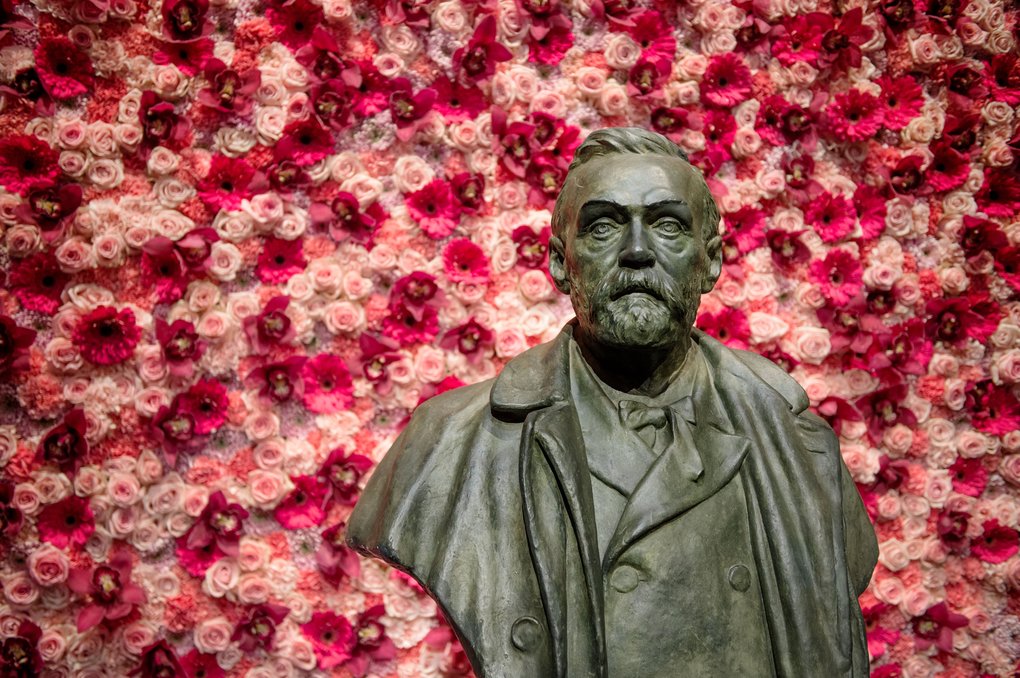 Bronsbyst av Alfred Nobel i skäggig och rock framför en vägg av rosa och röda blommor.