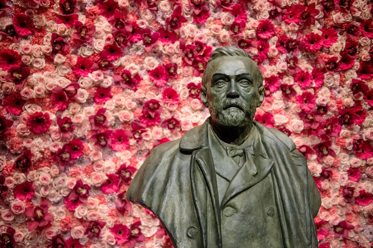 Bronsbyst av Alfred Nobel i skäggig och rock framför en vägg av rosa och röda blommor.