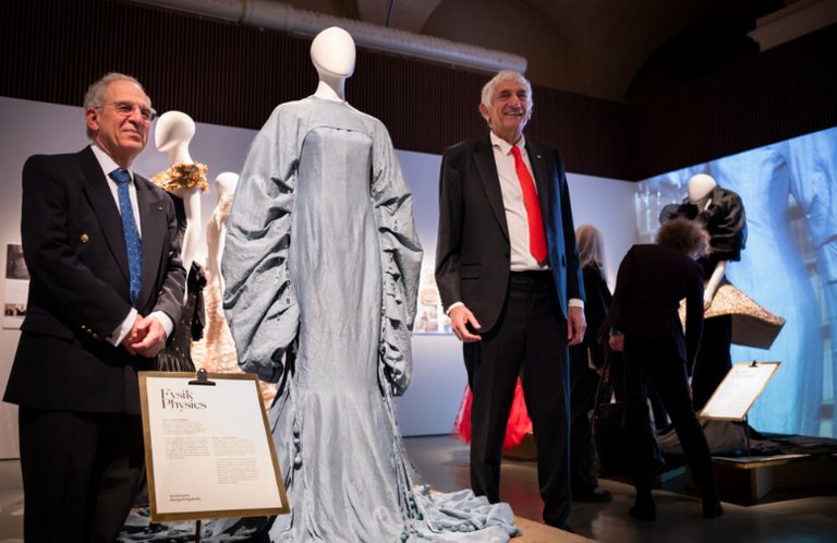 Two men in suits are standing next to a mannequin dressed in a grey, pleated gown at an exhibition. They hold informational boards and are in an art or fashion display with several mannequins in the background.