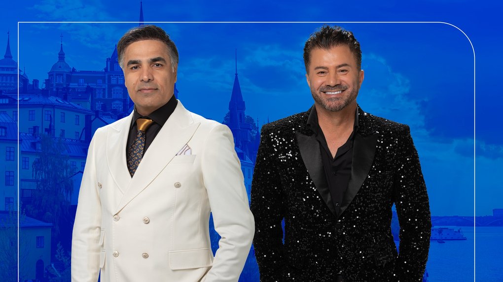 Two men pose in front of a blue background with a building. One is wearing a white suit and the other a black glittery jacket.