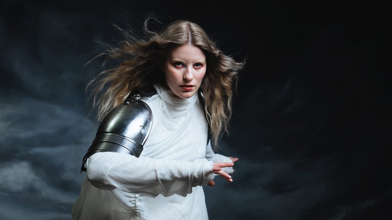A person with long blonde hair is wearing a white outfit and a metallic arm weapon, set against a dramatic background.