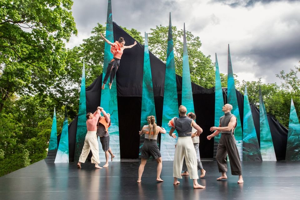 Actors and acrobats perform on an outdoor stage surrounded by tall, pointed set pieces in blue-green tones. One performer is thrown into the air while the others stand ready to catch them. The image conveys movement, teamwork, and dramatic stage performance.