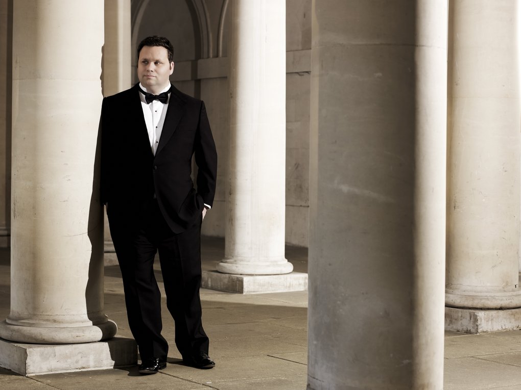 A man in black tie standing among marble columns.