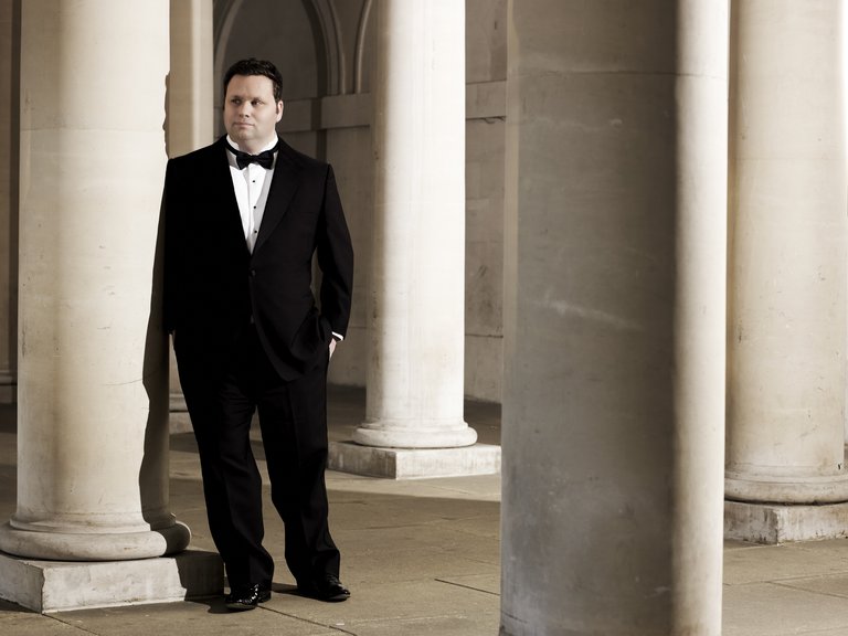 A man in black tie standing among marble columns.