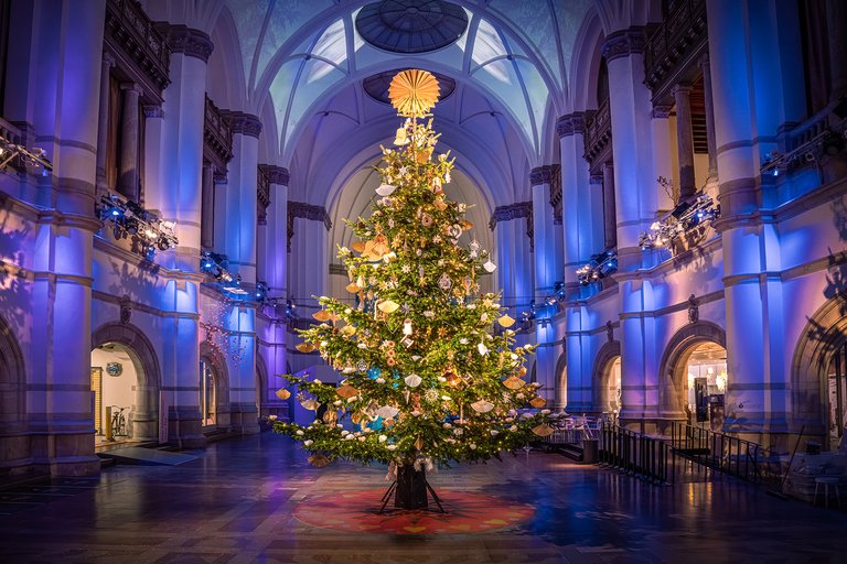 A big, well-decorated Christmas tree in the middle of a great hall.