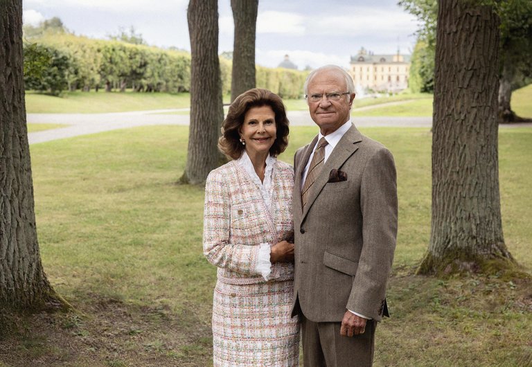 The King and Queen standing together in a park with trees and a palace in the background.