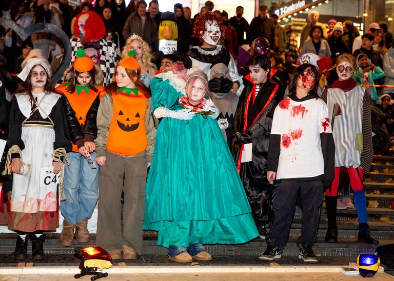A group is posing in various Halloween costumes on a stage with an audience in the background. The costumes include jack-o'-lantern themes, bloodstains, theatrical outfits, and horror masks.