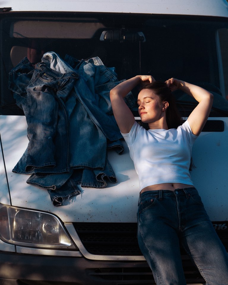Sigrid leaning back on the hood of a car with a bundle of denim pants beside her.