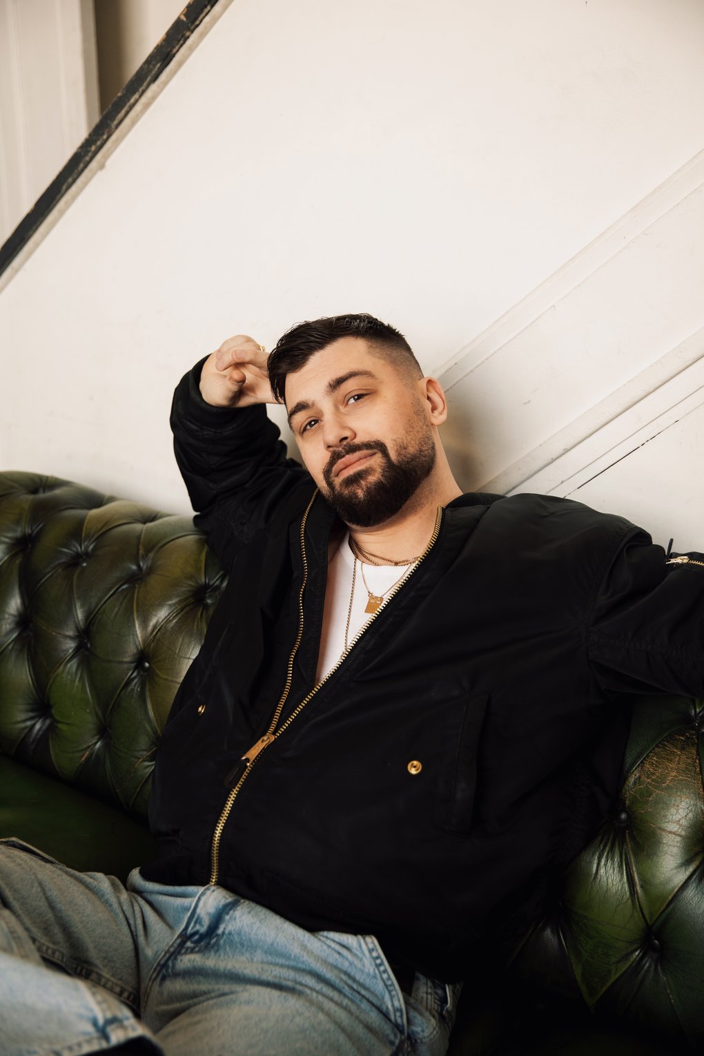 A relaxed man in jeans, a black jacket and a neat beard leaning back in a brown leather couch.