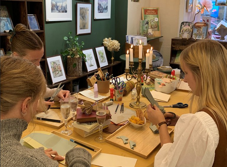 Three people are seated at a table working on craft or artistic projects. The table is covered with tools, materials, and small items such as scissors, brushes, and color samples.