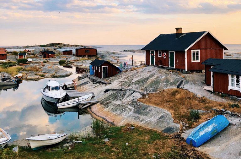 Stockholms sk'rgård. Huvudskär, sommar. Solnedgång. Vattnet ligger stilla mellan Huvudskärs kobbar. Flera röda fiskestugor syns på bilden, samt fiskebåtar.