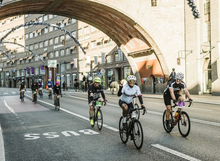 Cyclists compete in a group on an urban street under a decorated bridge with festive lights. The street is closed off and participants are wearing sports gear and helmets.