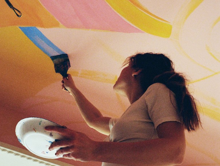 Artist painting a blue streak on a large pink ceiling.