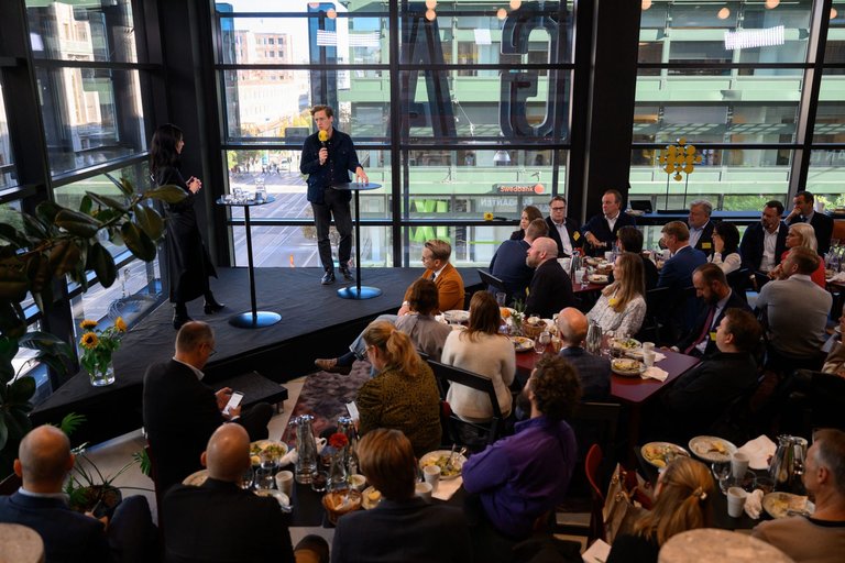A stage with two speakers before a crowd sitting at tables eating lunch