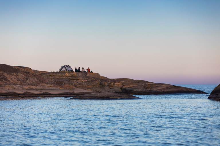 A group of four people sitting alongside a tent, on the cliffs of the island of Myggskar, surrounded by water.