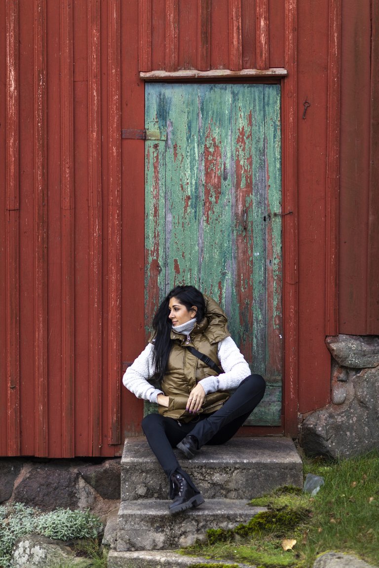 A woman with black hair is sitting in front of a red-painted wooden cottage.