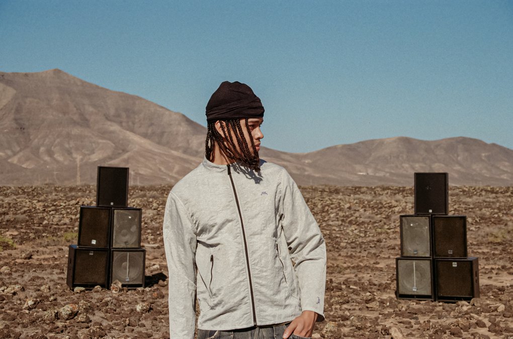 A man wearing a light gray jacket, standing in the desert in front of a mountain range with a speaker setup behind him.