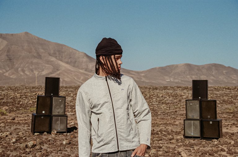 A man wearing a light gray jacket, standing in the desert in front of a mountain range with a speaker setup behind him.